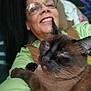 person, cat, siamese_cat, glasses, smiling, indoor, closeup, face, pet, cozy, nap, fur, whiskers, brown_cat, happy, human, arm, clothing, portrait, relaxing