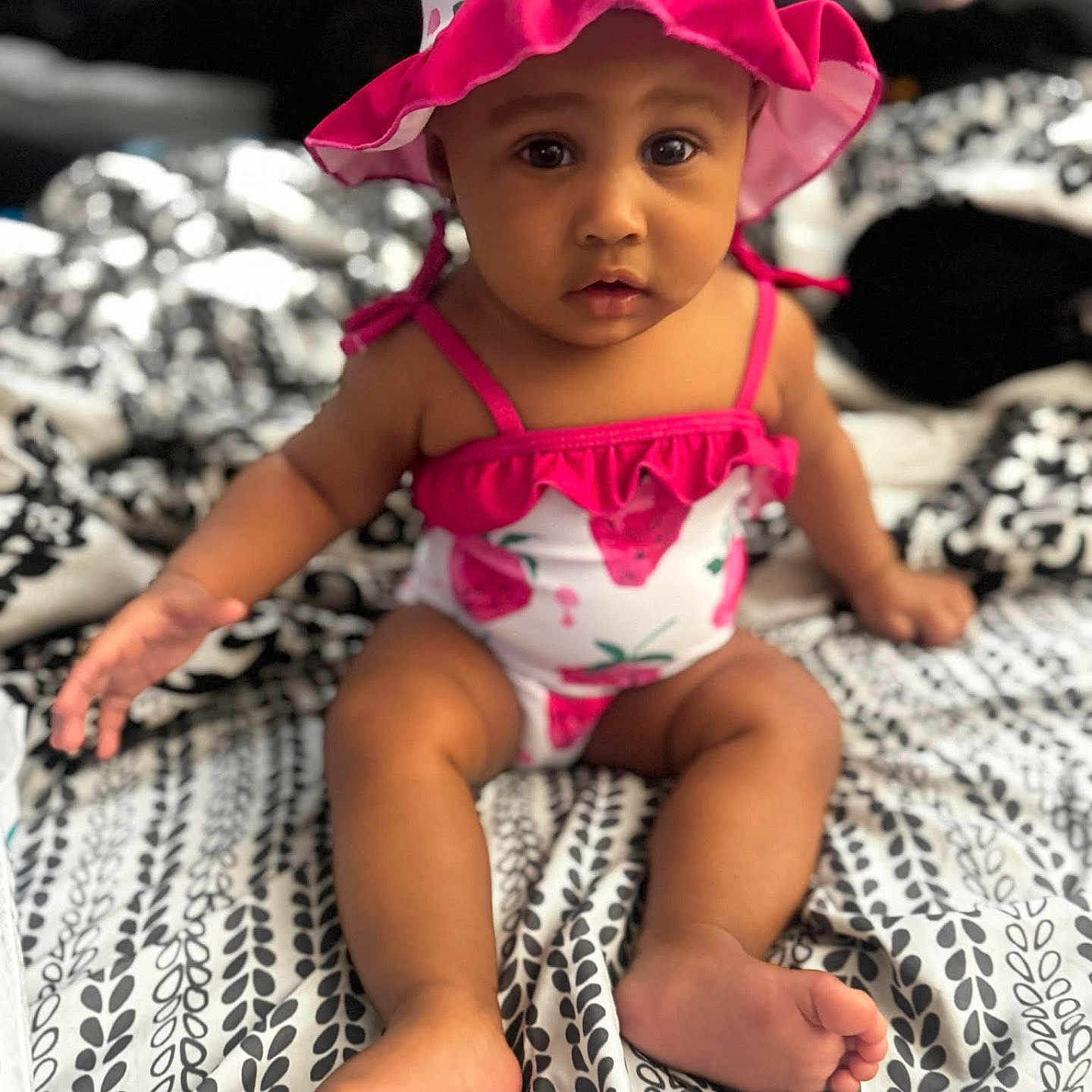 Faith is registered to the contest to win money with this photo: baby, infant, child, portrait, sitting, sun_hat, floral_clothing, pink_outfit, romper, bed, bedspread, indoors, face, eyes, serious_expression, barefoot, arms, legs, patterned_fabric, photography