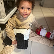 Gianni a rejoint le concours — aidez-le/la à gagner de superbes lots ! child, toddler, plush_toy, stuffed_animal, curly_hair, beige_clothing, floor_tiles, indoor, smile, happy, sitting, toy, person, home, cute, young_child, soft_toy, play, light, cozy