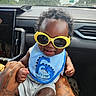 toddler, child, sunglasses, yellow, blue_bib, car_interior, hand_holding, tattoos, curly_hair, smile, rainy_window, dashboard, seat, clothing, person, portrait, happy, baby, cute, indoors