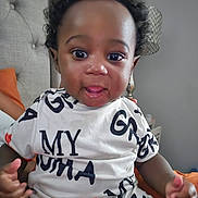 Syair Hayes is registered to the contest to win money with this photo: baby, child, curly_hair, black_hair, white_clothing, text_print, indoor, bedroom, headboard, lamp, person, sitting, cute, smiling, playful, young_child, portrait, skin, hand, furniture