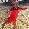 braids, car, child, crocs, daytime, footwear, girl, grass, happy, leaves, outdoor, person, playful, posing, red_bow, red_tracksuit, smiling, sunlight, vehicle, young