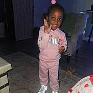Niyara Hayes joined the competition — help win amazing prizes! child, pink_tracksuit, smiling, indoor, living_room, cushioned_chair, wooden_cabinet, rug, shoes, flash_reflection, person, floor, hand_gesture, dark_background, home, toy, curtain, happy, casual_clothing, standing
