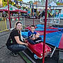 carnival, amusement_park, child, baby, woman, miniature_car, ride, outdoor, smiling, family, tent, lights, fence, asphalt, daytime, casual_clothing, playful, fun, people, portrait