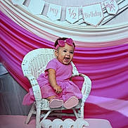 Sevyn is registered to the contest to win money with this photo: baby, child, birthday, portrait, smiling, sitting, pink_dress, hair_bow, wicker_chair, birthday_decorations, bunting, 2026, indoor, celebration, happy, infant, pink_background, studio_photo, dress, chair