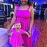 child, baby, mother, woman, smiling, happy, pink_dress, bow, sandals, standing, holding_hands, portrait, indoor, salon, neon_lighting, hair_salon, couch, wooden_floor, family, photography