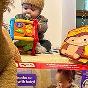 Elijah joined the competition — help win amazing prizes! baby, infant, pacifier, activity_cube, knit_hat, stuffed_animal, plush_toy, cloth_book, monkey_illustration, product_box, carpet, sitting, indoor, playtime, curious, hand, toy, colorful, portrait, toddler