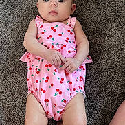 Elliana is registered to the contest to win money with this photo: baby, infant, child, pink_outfit, cherry_pattern, carpet, lying_down, hands, legs, skin, face, head, eyes, expression, indoor, cute, clothing, person, young, toddler