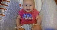 Elliana is registered to the contest to win money with this photo: baby, baby_seat, cheerful, child, clothing, comfort, cushion, face, happy, head, human, indoor, infant, looking_at_camera, person, red_shirt, seat_belt, sitting, smiling, young_child