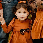 Harley is registered to the contest to win money with this photo: toddler, child, curly_hair, bow, holding_hands, orange_dress, black_bow, adult, jeans, casual_clothing, outdoor, portrait, family, cute, smiling, young_child, fall_colors, warm_light, close_up, standing