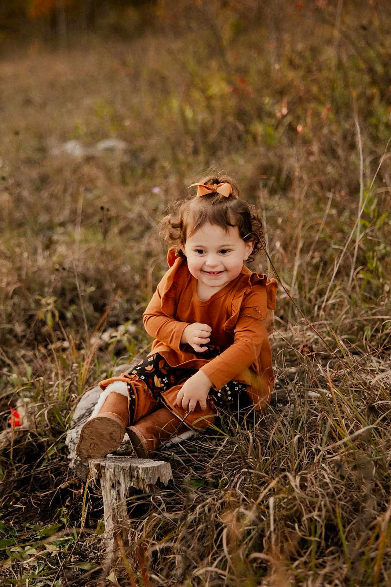 Harley is registered to the contest to win money with this photo: toddler, child, smiling, curly_hair, bow, orange_clothing, boots, sitting, tree_stump, grass, autumn, outdoor, nature, field, happy, young_child, cute, portrait, fall_colors, playful