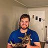 beard, blue_shirt, boxes, carpet, casual, cat, door, feline, happy, holding, human, indoor, man, pet, portrait, room, short_hair, smiling, tabby_cat, wall