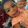 Yazmin is registered to the contest to win money with this photo: toddler, child, smiling, waving, face, eyes, mouth, teeth, hair, peach_dress, patterned_clothing, blanket, tie_dye, fabric, indoor, portrait, happy, cute, person, closeup