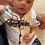 baby, child, infant, person, clothing, baseball_jersey, shorts, gold_chain, jewelry, sitting, curly_hair, skin, hand, lap, indoor, portrait, casual, black_and_white, cute, young