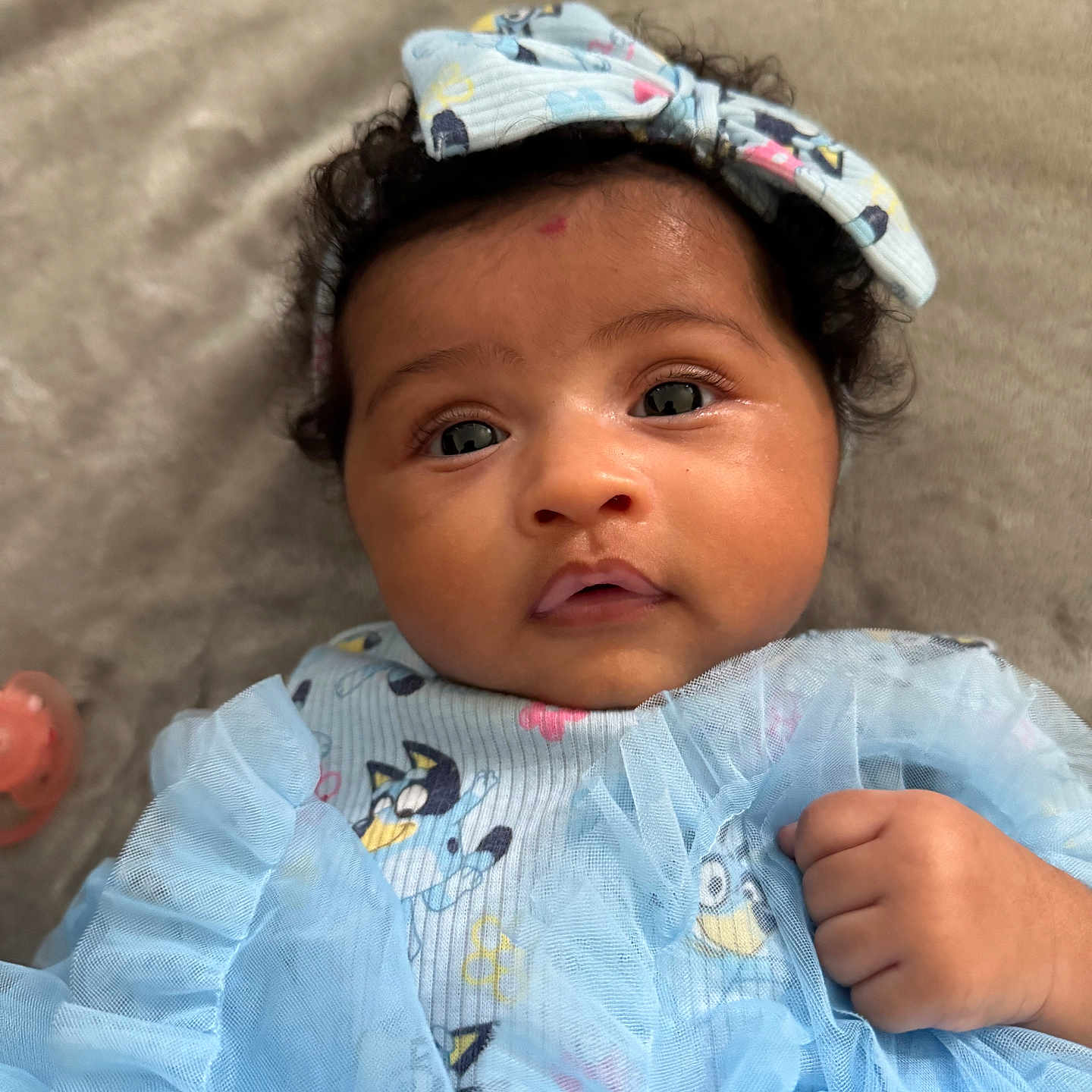 Novah is registered to the contest to win money with this photo: baby, child, infant, portrait, close_up, face, eyes, bow, headband, blue_outfit, dress, ruffles, curly_hair, lying_down, indoors, blanket, adorable, wide_eyed, hand, photography