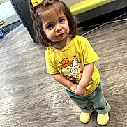 Jasminemiamor Bitter is registered to the contest to win money with this photo: toddler, child, yellow_bow, yellow_shirt, cartoon_print, curious_look, indoor, wooden_floor, green_pants, yellow_shoes, modern_furniture, bench, flooring, portrait, standing, cute, young_child, casual_clothing, small_person, hands_clasped