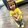 toddler, child, yellow_bow, yellow_shirt, cartoon_print, curious_look, indoor, wooden_floor, green_pants, yellow_shoes, modern_furniture, bench, flooring, portrait, standing, cute, young_child, casual_clothing, small_person, hands_clasped