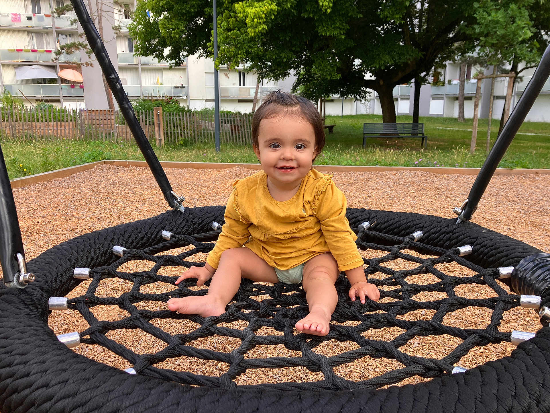 Ines participe au concours pour gagner de l'argent avec cette photo : toddler, child, playground, swing, rope_swing, smiling, barefoot, yellow_shirt, outdoor, grass, tree, bench, wood_chips, residential_building, happy, person, daylight, park, seat, nature