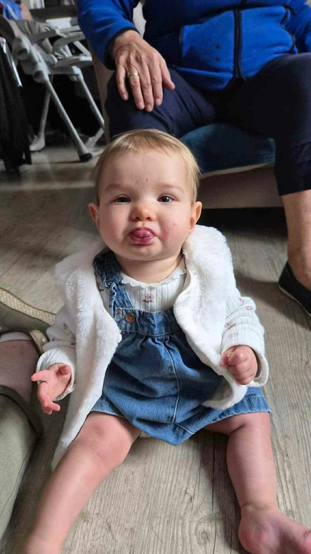 Inaya a rejoint le concours — aidez-le/la à gagner de superbes lots ! baby, child, tongue, denim_dress, white_jacket, sitting, floor, wooden_floor, hand, adult, indoor, cute, person, clothing, legs, face, expression, playful, casual, home