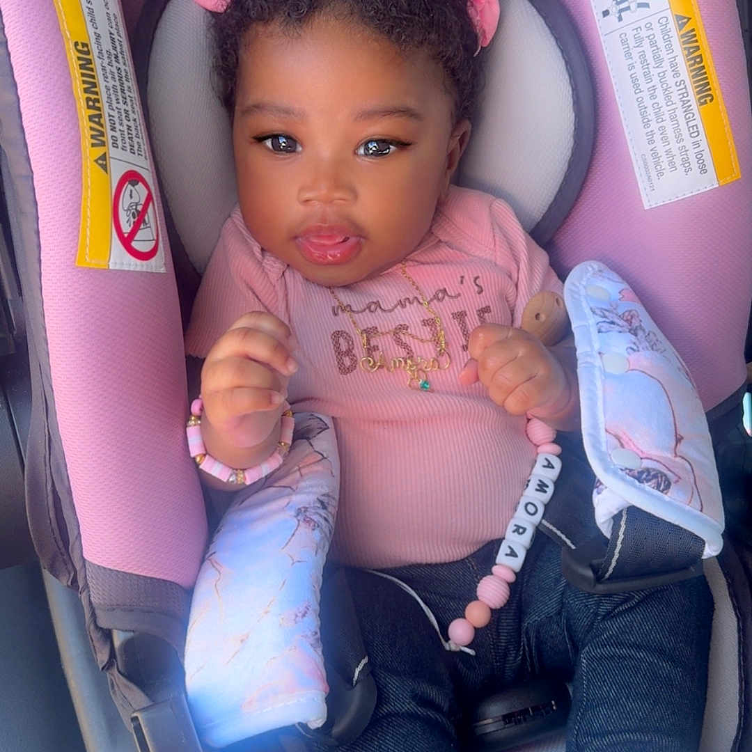 Amora is registered to the contest to win money with this photo: baby, child, car_seat, pink, clothing, hair_bow, hair_clip, denim, shoes, bracelet, necklace, infant, cute, portrait, indoor, seat_belt, accessory, person, smiling, young