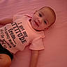 Maelyn is registered to the contest to win money with this photo: baby, infant, child, portrait, smiling, lying_down, bed, pink, clothing, shirt, text_on_clothing, indoors, bedroom, close_up, face, eyes, happy, casual, photography, home