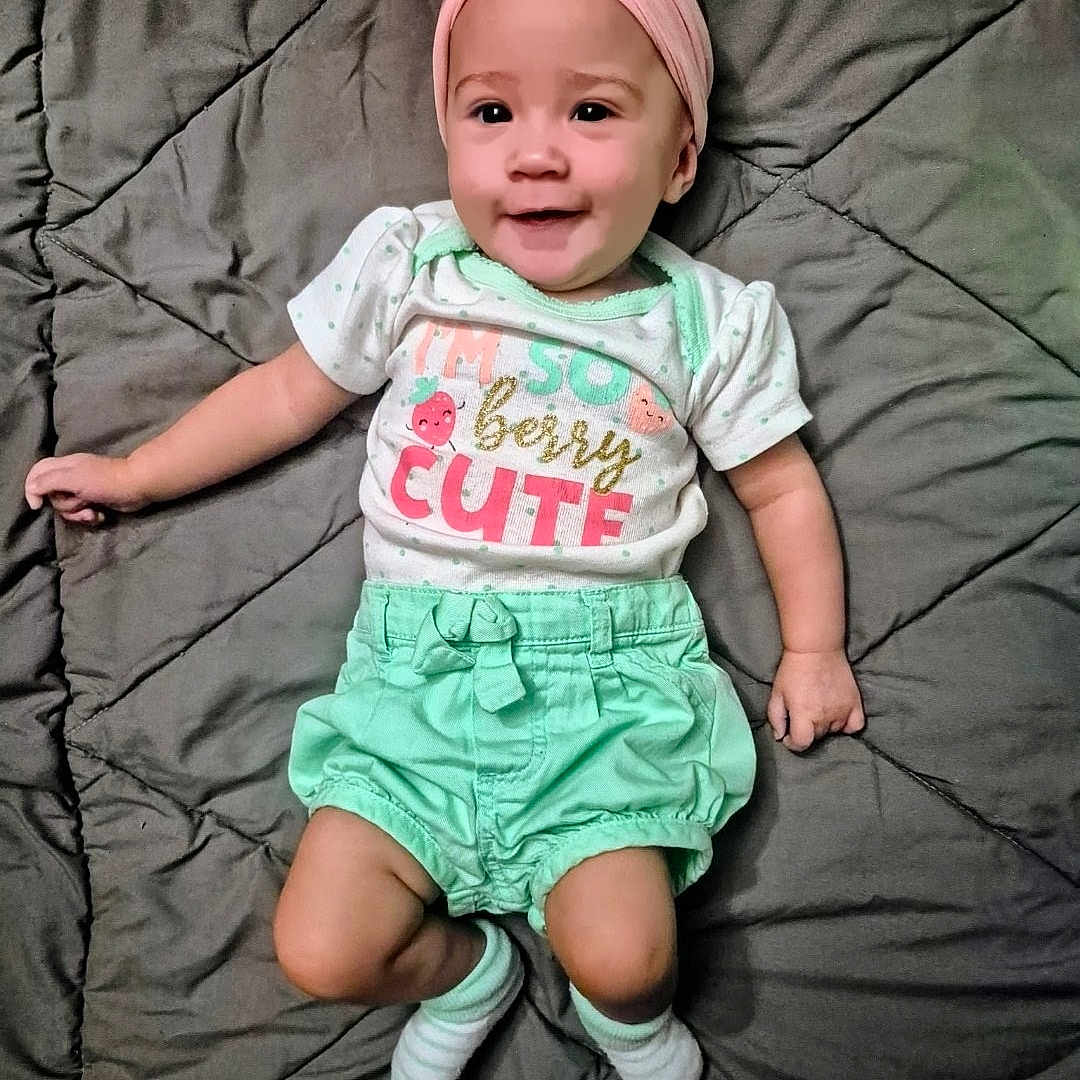Maelyn is registered to the contest to win money with this photo: baby, child, infant, smiling, portrait, lying_down, bed, quilt, bow, headband, pink, mint_green, clothing, t_shirt, shorts, socks, cute, indoors, happy, photography