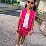 child, girl, outdoor, pink_clothing, shorts, vest, sunglasses, heart_shaped, hair_accessory, bracelet, watch, sandals, standing, urban, concrete, daylight, fashion, serious_expression, person, summer