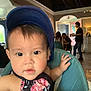 baby, blue_cap, casual, chair, child, curious, dining, face, floral_outfit, hand, hat, high_chair, indoor, light, painting, people, person, restaurant, table, wall