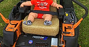 Jax joined the competition — help win amazing prizes! baby, child, lawn_mower, orange, greenery, grass, outdoor, seat, smiling, red_shirt, socks, safety_belt, nature, cute, happy, person, summer, machine, equipment, park