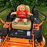 baby, child, lawn_mower, orange, greenery, grass, outdoor, seat, smiling, red_shirt, socks, safety_belt, nature, cute, happy, person, summer, machine, equipment, park