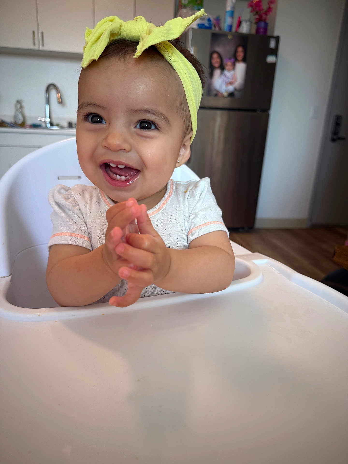 Araceli joined the competition — help win amazing prizes! toddler, child, high_chair, yellow_headband, clapping, smiling, kitchen, refrigerator, family_photo, white_shirt, indoor, person, face, happy, baby, furniture, wood_floor, cabinet, sink, home