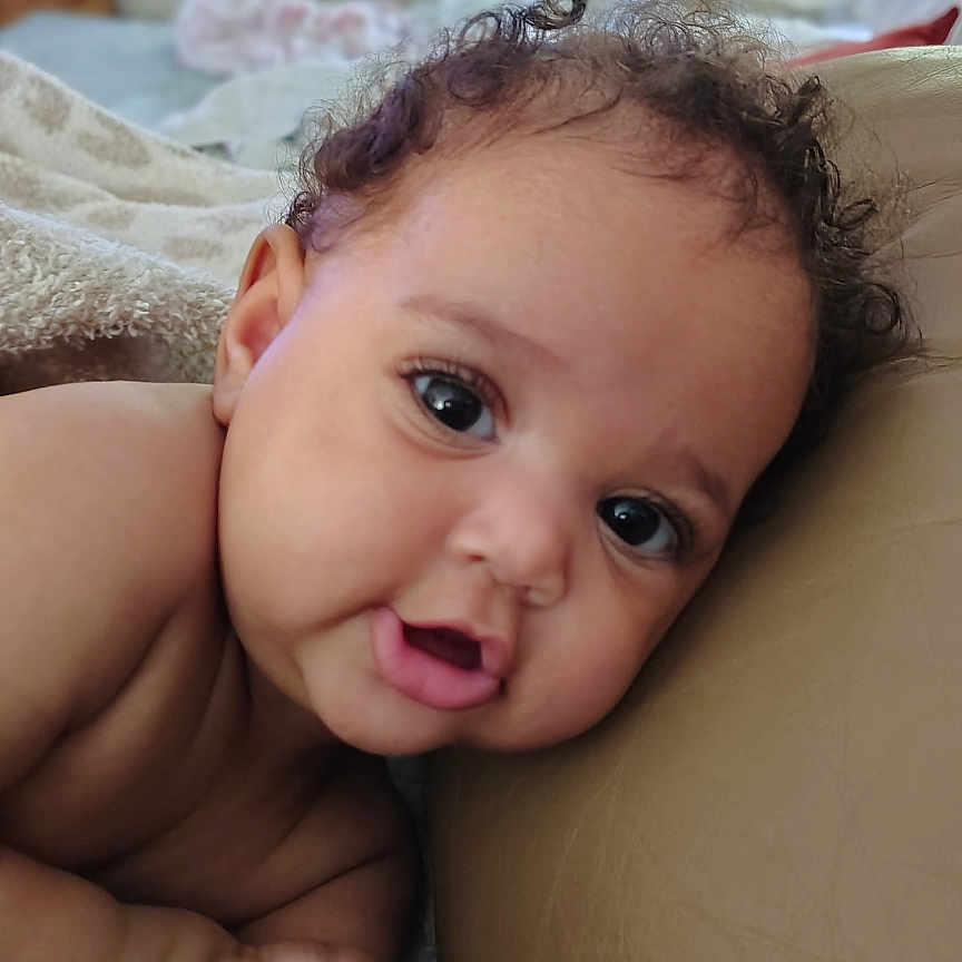 Gianna is registered to the contest to win money with this photo: baby, infant, face, eyes, curly_hair, skin, arm, hand, couch, indoor, soft_light, expression, portrait, closeup, child, resting, cute, natural_light, home, cozy