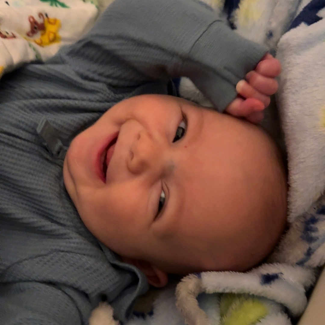 Leroy is registered to the contest to win money with this photo: baby, blanket, child, colorful, comfort, cozy, cute, expression, face, happy, indoors, infant, lying_down, playful, portrait, sleepwear, smiling, soft, toddler, warm