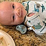 Leroy is registered to the contest to win money with this photo: baby, infant, smiling, face, pacifier, clothing, dinosaur_pattern, granite_countertop, kitchen, plate, plastic_spoon, person, child, cute, lying_down, indoors, happy, closeup, baby_clothes, beads