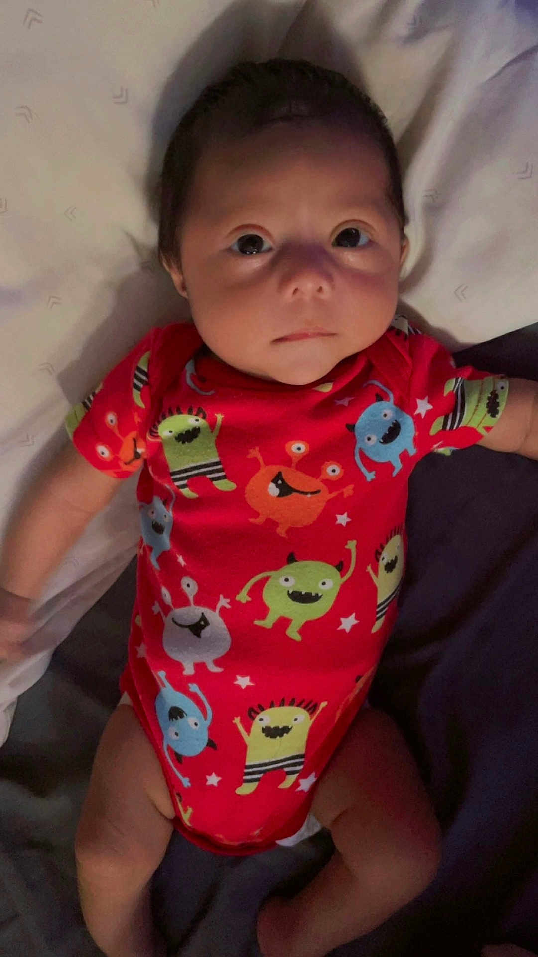 Jose Antonio is registered to the contest to win money with this photo: baby, infant, onesie, red_clothing, cartoon_monsters, bed, blanket, lying_down, cute, child, face, looking_at_camera, soft_lighting, indoor, skin, head, arms, legs, newborn, expressionless