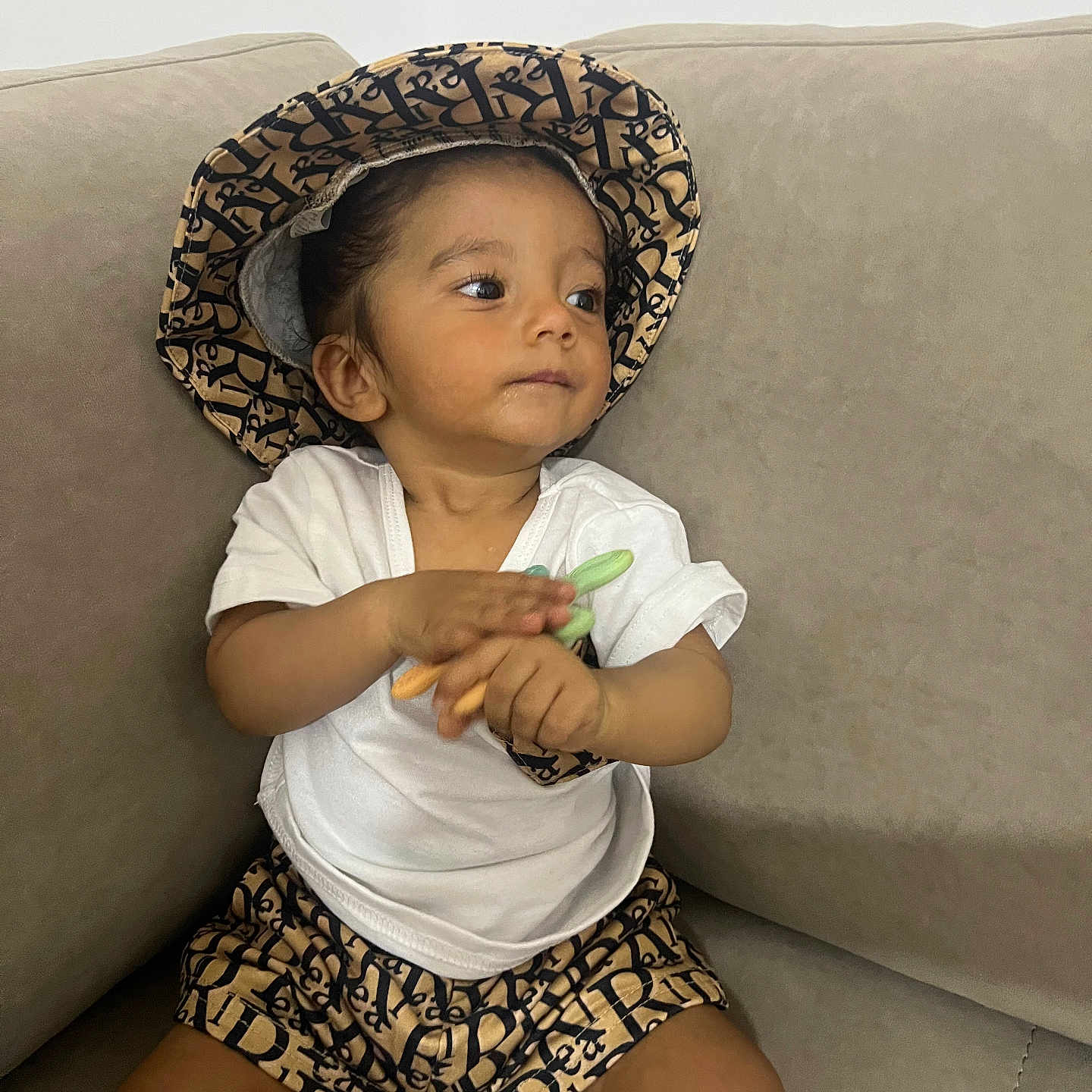 Melwan participe au concours pour gagner de l'argent avec cette photo : beige, brown_skin, casual_clothing, child, clapping, comfortable, couch, curious, hat, indoor, patterned_clothing, portrait, relaxed, short_hair, shorts, sitting, toddler, toy, white_tshirt, young_child