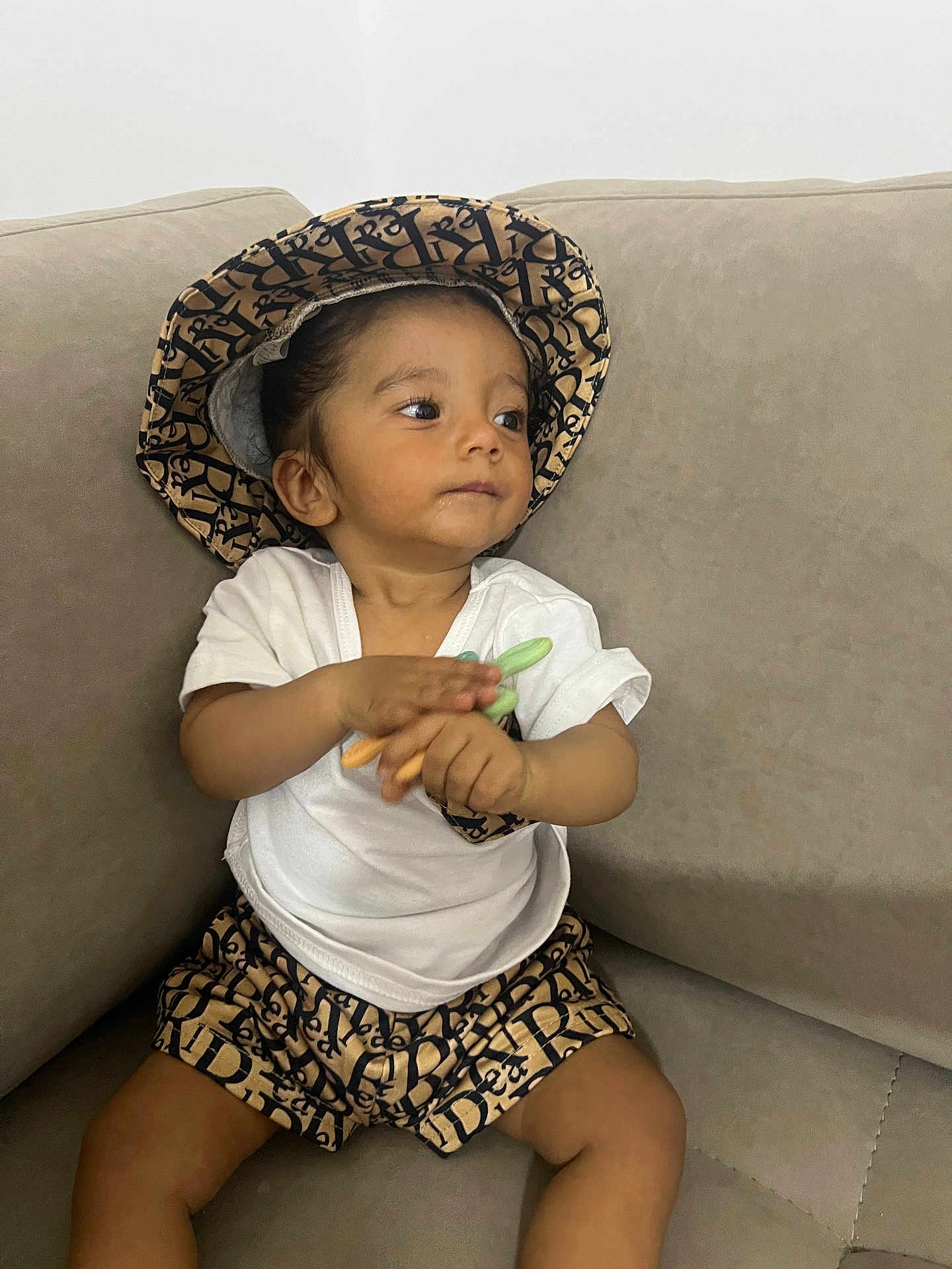 Melwan participe au concours pour gagner de l'argent avec cette photo : toddler, child, hat, shorts, white_tshirt, couch, beige, clapping, toy, indoor, casual_clothing, patterned_clothing, portrait, young_child, sitting, curious, brown_skin, short_hair, comfortable, relaxed