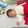 baby, sleeping, infant, bed, blanket, plush_toy, stuffed_animal, blue_toy, red_blanket, white_clothing, portrait, indoors, bedroom, peaceful, resting, face, closed_eyes, hand, bedding, cozy
