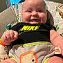 baby, infant, child, smiling, chubby_cheeks, nike, logo, onesie, baby_bouncer, baby_seat, colorful_pattern, toy, hands, legs, window, blinds, indoor, portrait, closeup, happy
