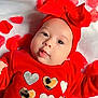 baby, infant, red_clothing, heart_pattern, headband, bow, rose_petals, blanket, portrait, cute, child, smiling, face, soft_texture, lying_down, closeup, adorable, festive, warm_colors, jewelry