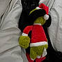 black_cat, cat, plush_toy, grinch, stuffed_animal, bed, blanket, white_bedding, cute, cozy, holiday, christmas, festive, animal, pet, claws, soft_texture, indoor, sleepy, hugging