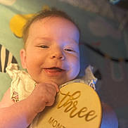 Aerilyn is registered to the contest to win money with this photo: baby, background, child, closeup, clothing, cute, face, hand, happy, indoor, infant, person, portrait, skin, smile, soft_light, text, three_months, wooden_plaque, young
