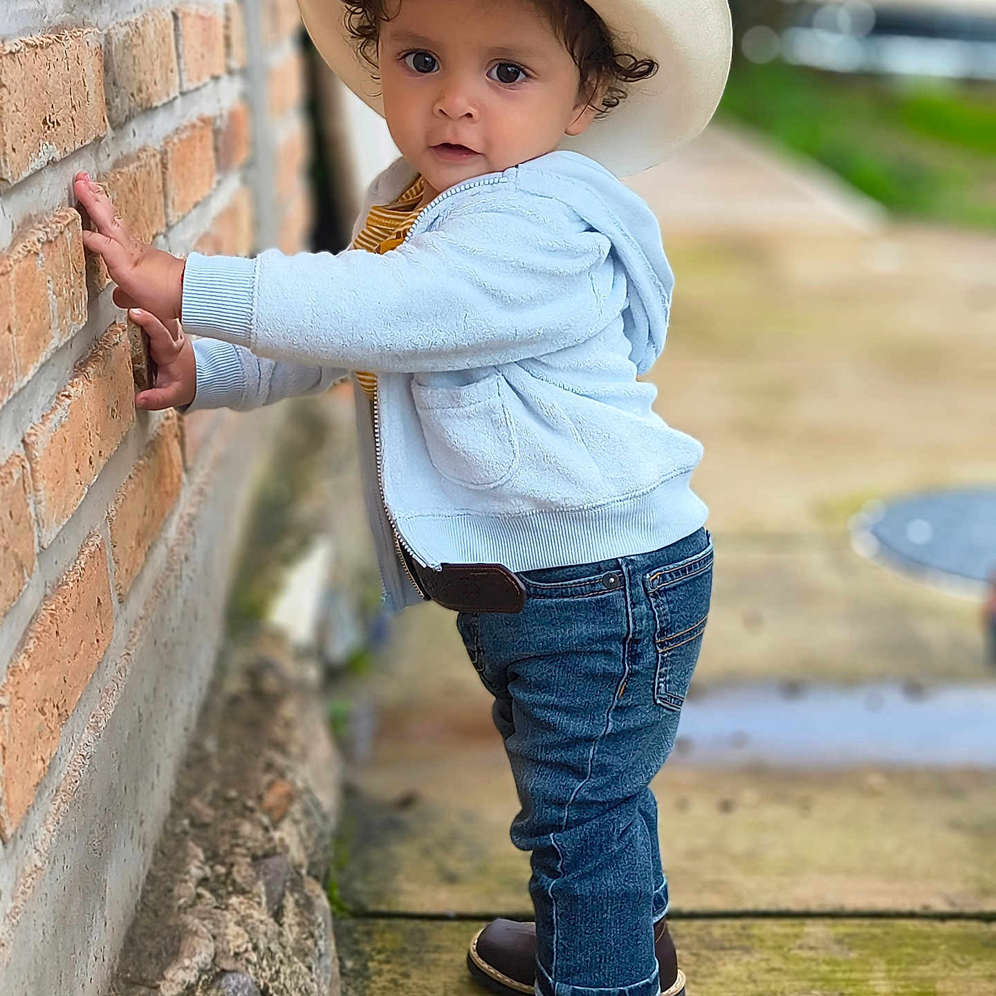 Ismael is registered to the contest to win money with this photo: boots, brick_wall, brown_eyes, casual_clothing, child, cowboy_hat, curious, curly_hair, cute, daylight, hoodie, jeans, outdoor, portrait, sidewalk, small_person, standing, toddler, urban, young_child