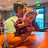 arm, baby, background, casual_clothing, child, curious, exit_sign, face, fingers, hair, holding, indoor, lighting, man, person, restaurant, seat, table, window, wooden_table