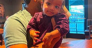 Lakshmi is registered to the contest to win money with this photo: arm, baby, casual_clothing, child, curious, exit_sign, facial_expression, holding, indoor, light, man, person, purple_clothing, restaurant, seated, shadow, short_hair, table, window, wood