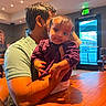 arm, baby, casual_clothing, child, curious, exit_sign, facial_expression, holding, indoor, light, man, person, purple_clothing, restaurant, seated, shadow, short_hair, table, window, wood