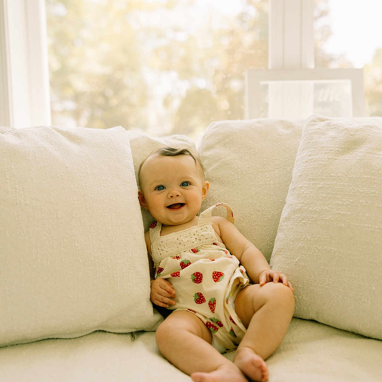 Oaklynn is registered to the contest to win money with this photo: baby, child, clothing, couch, cozy, cute, feet, happy, home, indoor, infant, legs, natural_light, pillow, portrait, sitting, skin, smiling, strawberry_pattern, window