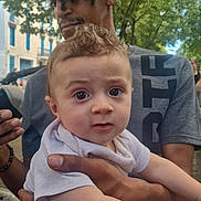 Mérého a rejoint le concours — aidez-le/la à gagner de superbes lots ! baby, man, outdoor, trees, cap, holding, person, curious, wide_eyes, casual_clothing, daylight, portrait, expression, nature, hand, bench, urban, street, people, family