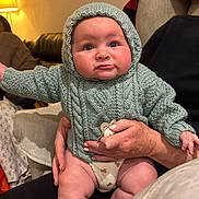 Llewelyn is registered to the contest to win money with this photo: baby, knitted_hoodie, indoor, couch, adult_hand, warm_lighting, close_up, cute, child, person, seated, cozy, expression, infant, home, soft_furniture, casual, portrait, human, warm_colors