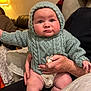 baby, knitted_hoodie, indoor, couch, adult_hand, warm_lighting, close_up, cute, child, person, seated, cozy, expression, infant, home, soft_furniture, casual, portrait, human, warm_colors